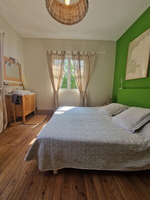 Quarto com piso de madeira, cama com colcha listrada cinza, cômoda de madeira, espelho e janela com cortinas.