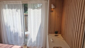 A bedroom with a wooden floor, a bed with a pink blanket, a small table with a lamp, and a sliding glass door leading to an outdoor area.