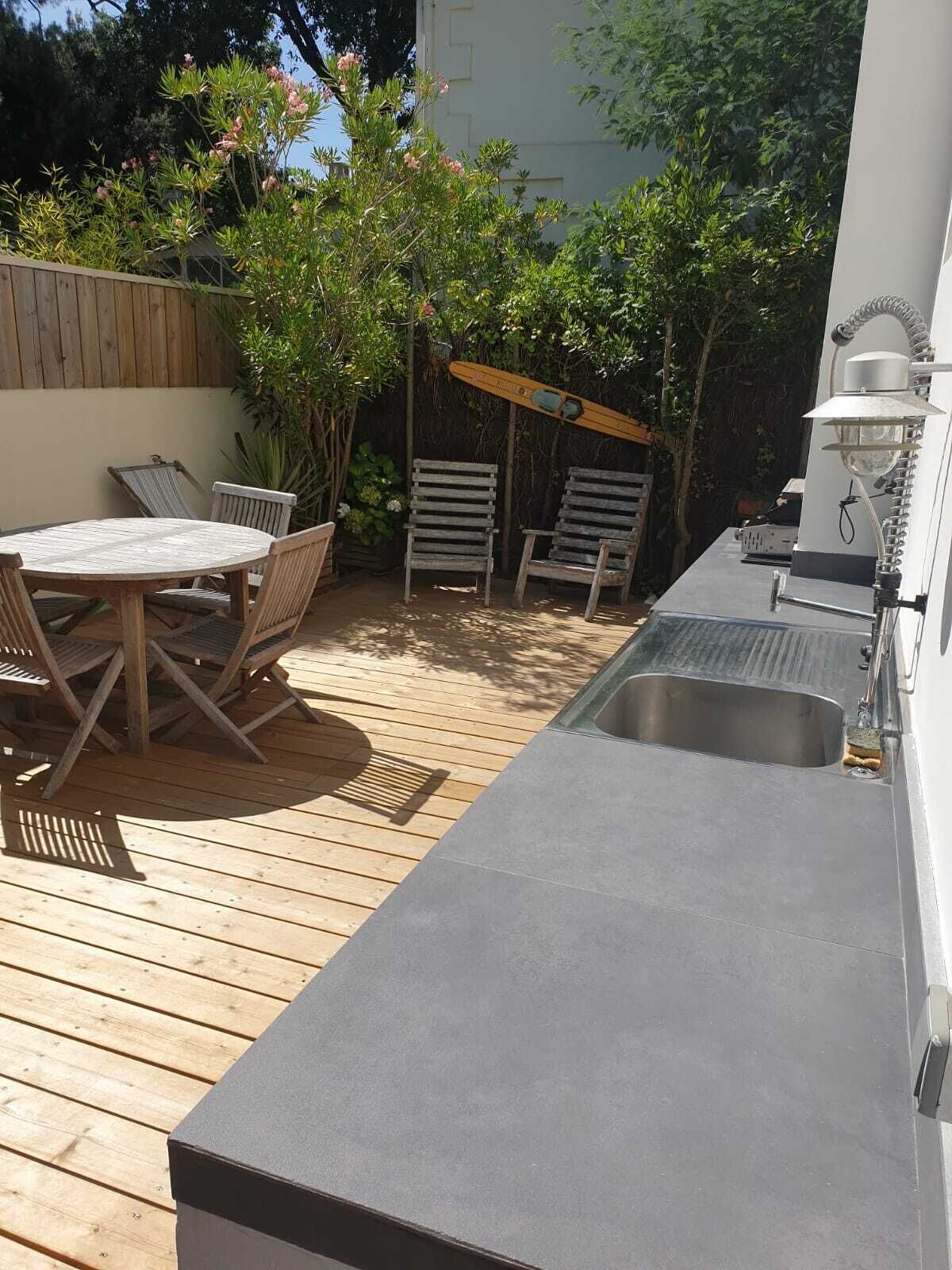 A wooden deck with a round table and chairs, a built-in sink, and a small garden area with plants and a pergola.