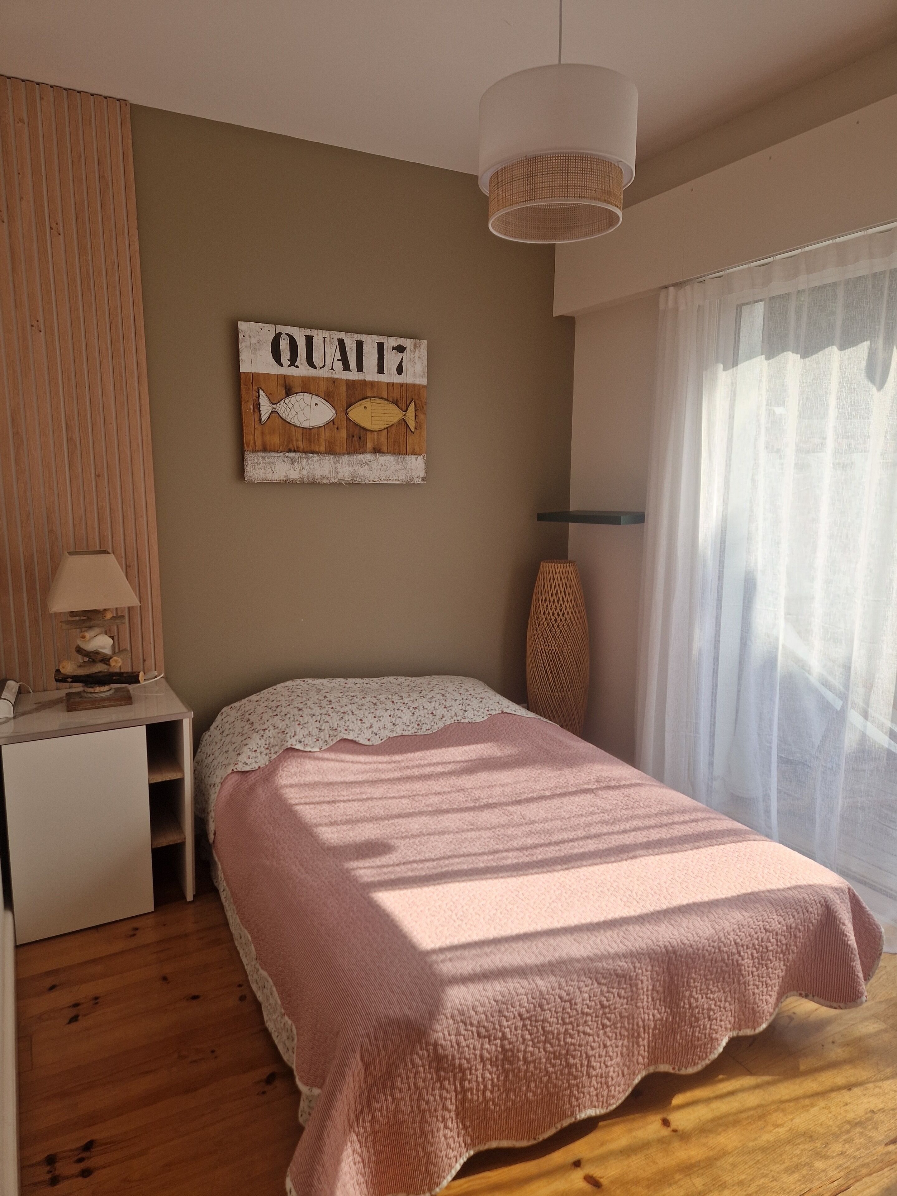Ein Schlafzimmer mit Bett, Nachttisch, Lampe, Wandbild und einem Fenster mit Vorhängen.