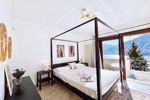 A four-poster bed with a view of mountains, a small bedside table with a lamp, and a framed picture on the wall. - Villa Isola Verde, Valsolda, Italy (Valsolda)