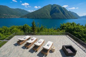 A terrace with lounge chairs and a table, overlooking a lake and mountains. - Villa Isola Verde, Valsolda, Italy (Valsolda)