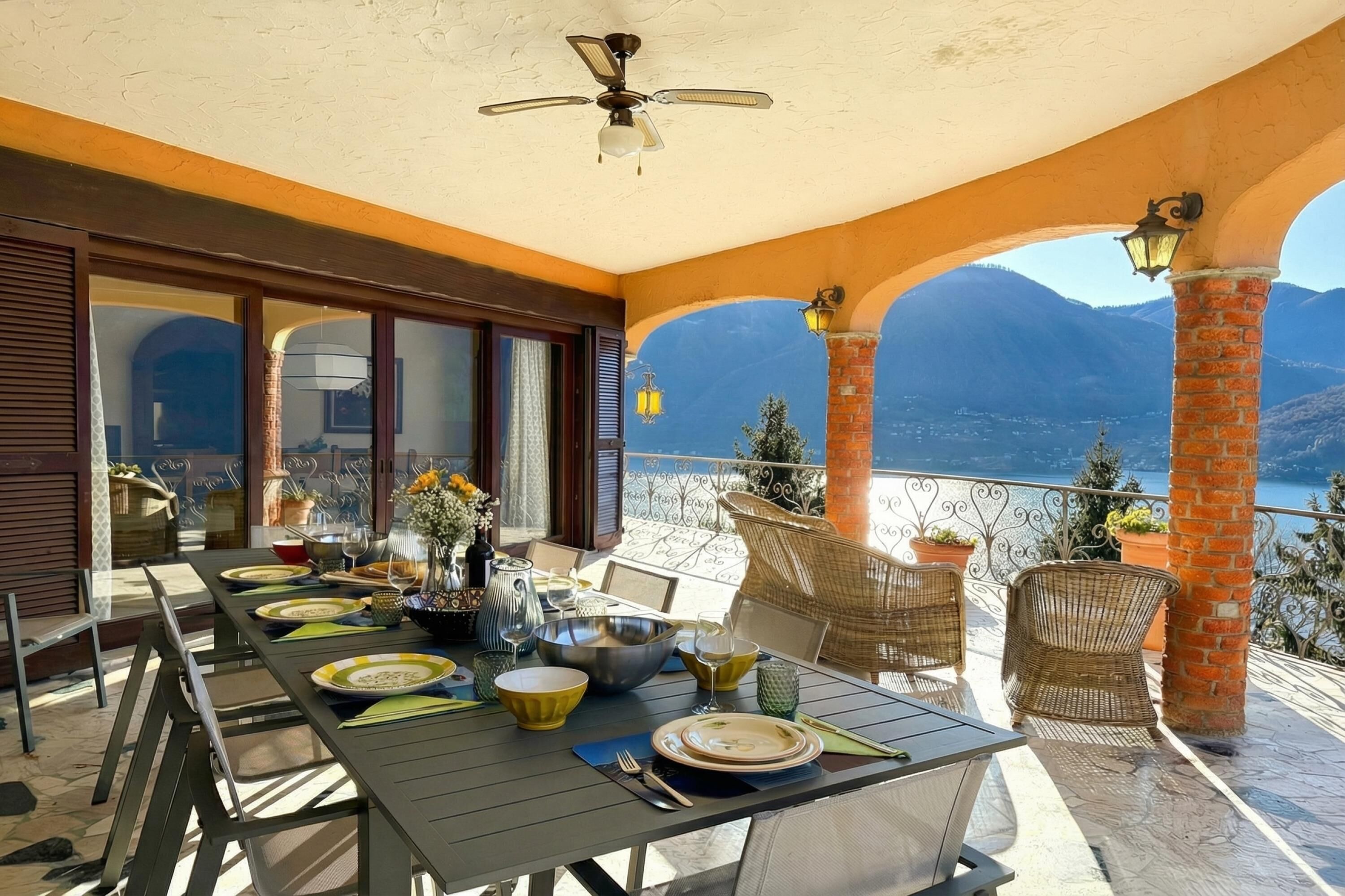 Une terrasse avec une table dressée pour le repas, des chaises en osier, un ventilateur de plafond et une vue sur les montagnes.