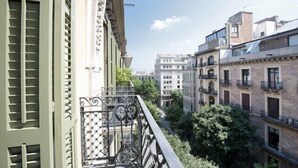 Una calle urbana estrecha flanqueada por edificios, con balcones de rejas de hierro forjado y contraventanas.