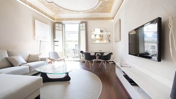 Una sala moderna con sofá, una mesa de centro de vidrio, un comedor con sillas, un televisor montado en la pared y un balcón visible a través de la puerta abierta.