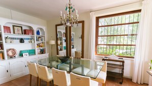 A dining area with a glass table, four chairs, a chandelier, a large window with a view of greenery, and a white cabinet with various items. - Wonderful Villa, private pool, seafront, garden, excellent views to the bay (Playa Albufereta- Alicante)
