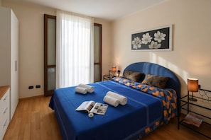 Une chambre d’hôtel comprenant un lit, une table de chevet, une lampe, une fenêtre avec des rideaux, une porte et un tableau accroché au mur.