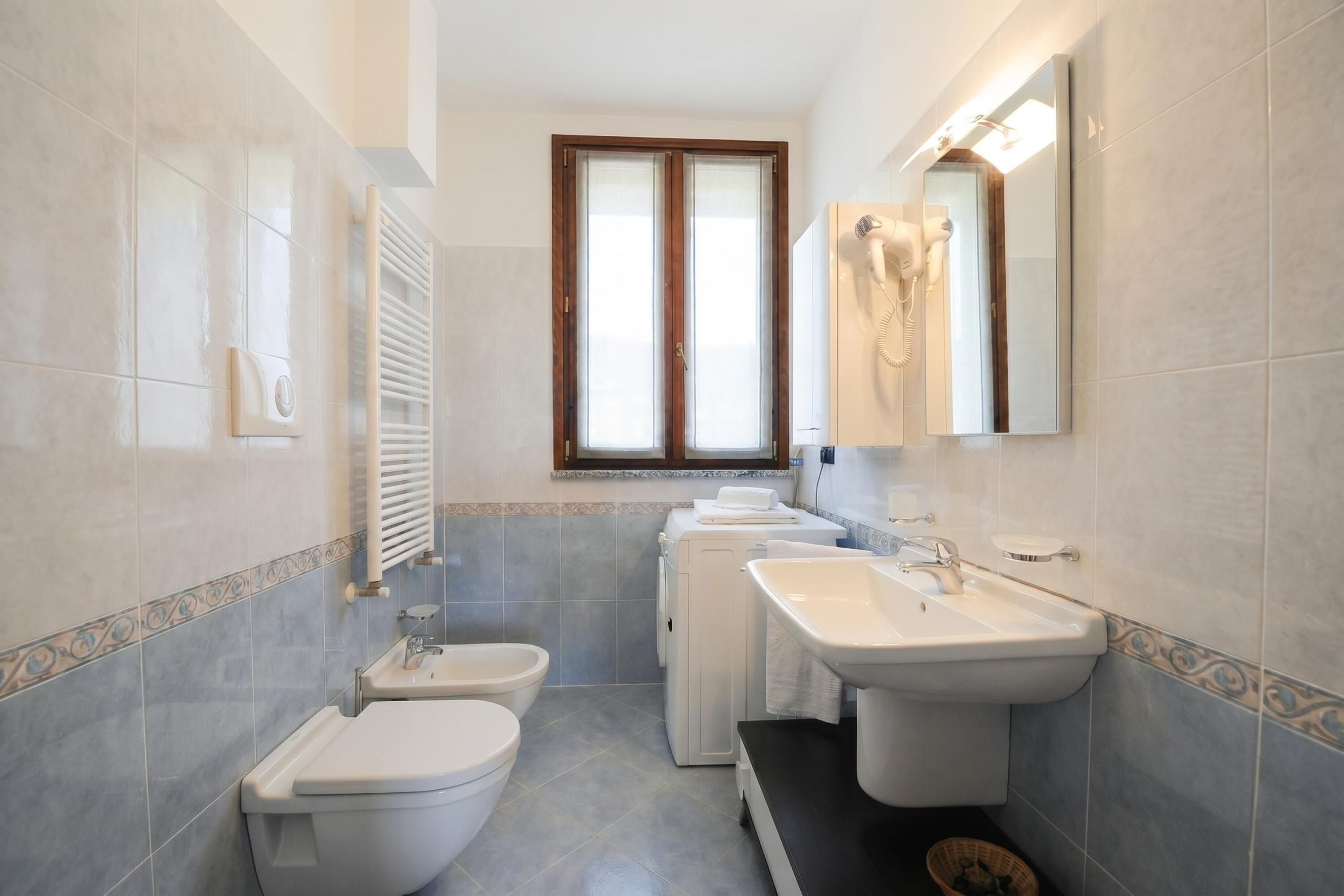 A bathroom with a toilet, bidet, two sinks, a hairdryer, and a window.