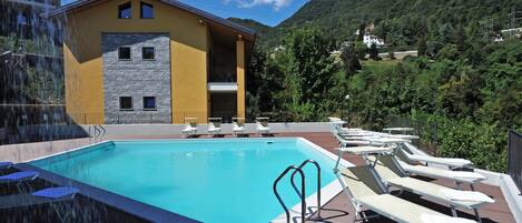 Una piscina con sillas de descanso, un edificio de fachada de piedra y un paisaje montañoso de fondo.