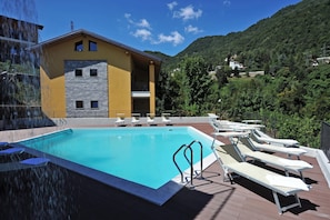 A swimming pool with lounge chairs, a building with a stone facade, and a mountainous backdrop. - Residence Eden Gardenia 11, Argegno, Italy (Argegno)