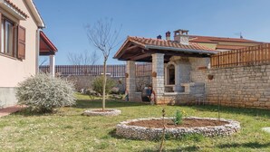 Une maison en pierre avec un toit en tuiles, un petit jardin avec un arbre et un parterre de fleurs, et une clôture en bois.