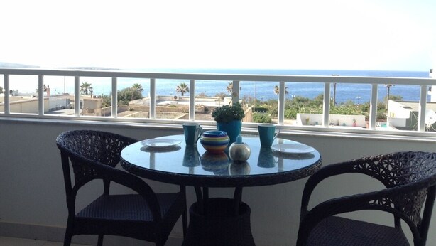 A balcony with a round table and two chairs, overlooking a coastal view with buildings and palm trees. - LUXURY SEAFRONT APARTMENT WITH POOL (Saint Paul's Bay)