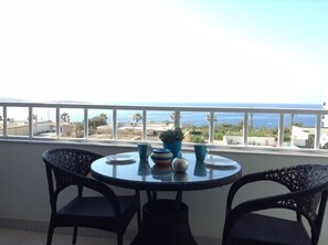 Un balcon avec une table ronde et deux chaises, offrant une vue sur la côte, avec des bâtiments et des palmiers.