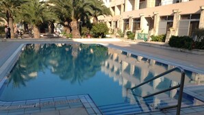A swimming pool with a clear blue water surface, surrounded by palm trees and a building with a beige facade. - LUXURY SEAFRONT APARTMENT WITH POOL (Saint Paul's Bay)