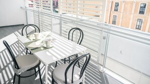 Un balcone con tavolo e sedie bianche, una ringhiera bianca e vista sugli edifici sullo sfondo.