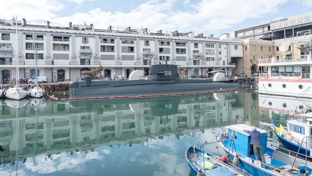 Un porto con una grande nave militare ormeggiata, un edificio con numerosi balconi e una barca più piccola in primo piano.