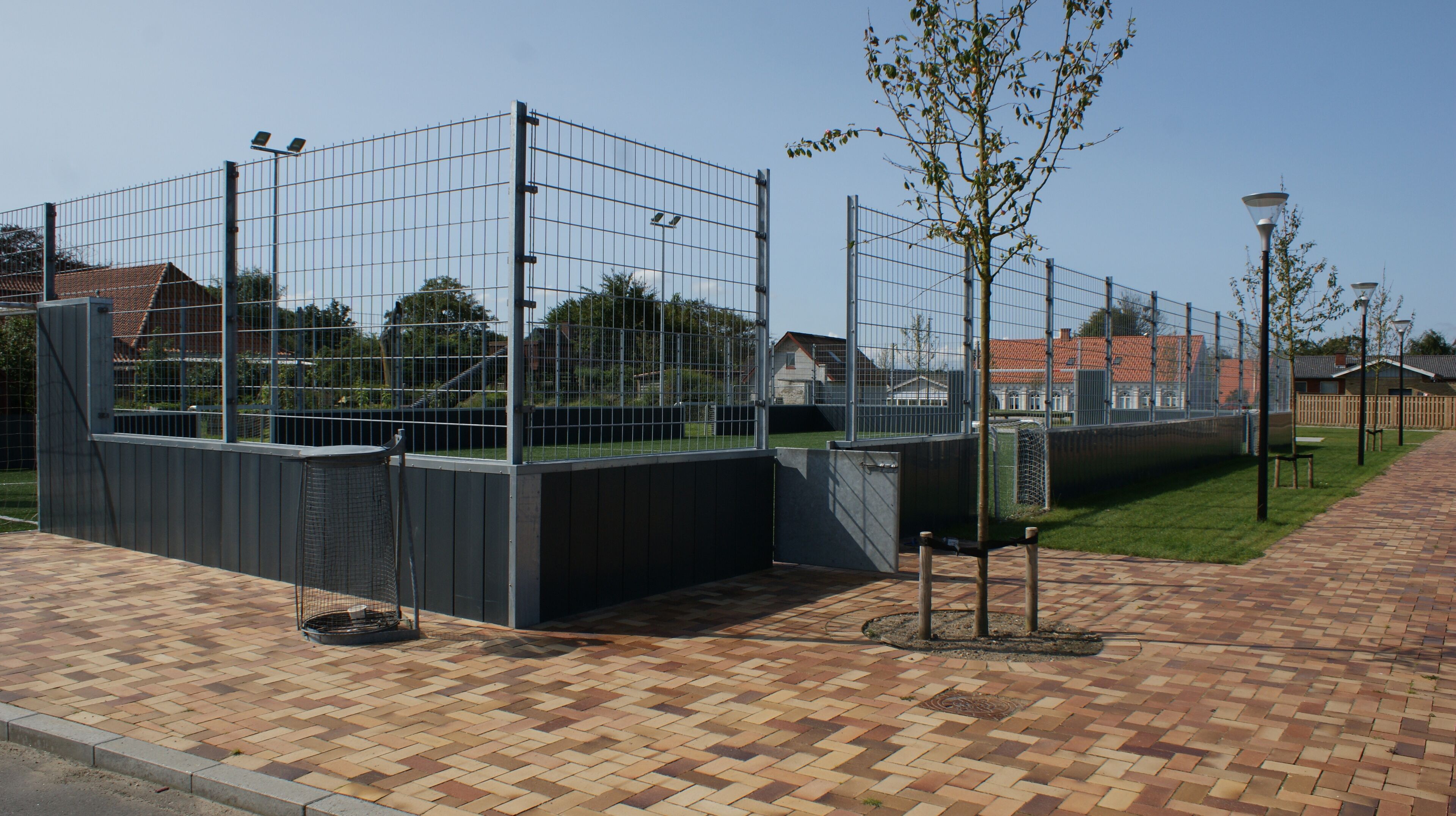 A fenced sports facility with a paved walkway, a small tree, and a lamp post.