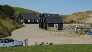 A gravel path leading to a row of black buildings with sloped roofs, a car parked in the foreground, and a rocky shoreline with boats in the background. - Wonderfull holiday house                                           (Nykøbing Mors)