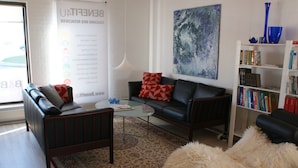 A modern living room with a black leather sofa, a glass coffee table, and a patterned rug. There is a bookshelf filled with books and decorative items. - Wonderfull holiday house                                           (Nykøbing Mors)