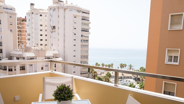 Ein Balkon mit Tisch und Stühlen, einem Blumentopf und Blick auf das Meer und Gebäude.