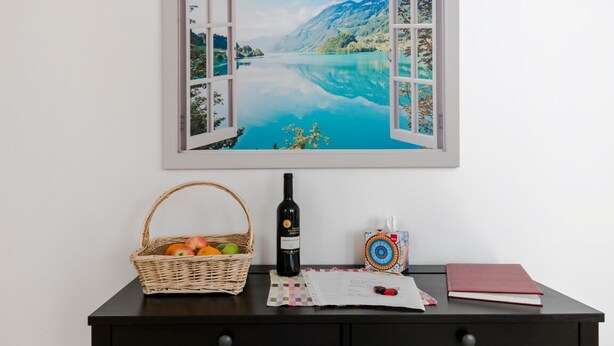 A wall-mounted artwork depicting a scenic lake with mountains, a wicker basket with fruits, a wine bottle, a notebook, and a red book on a dark wooden table. - Luxury newly renovated 3-bedroom, 2-bathroom mews house in Hampstead London (London)
