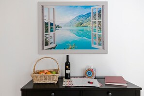 Wanddecoratie toont een schilderachtig meer met bergen, een rieten mand met fruit, een fles wijn, een notitieboekje, en een rood boek op een donkerhouten tafel.