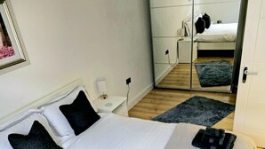 A neatly made bed with white linens and two dark pillows, a bedside table with a lamp, and a large mirror reflecting the room. - Luxury newly renovated 3-bedroom, 2-bathroom mews house in Hampstead London (London)