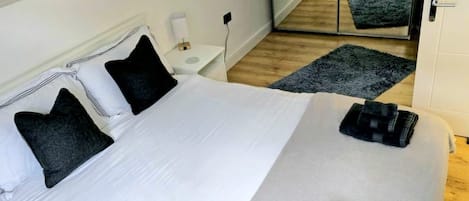 A neatly made bed with white linens and two dark pillows, a bedside table with a lamp, and a large mirror reflecting the room.