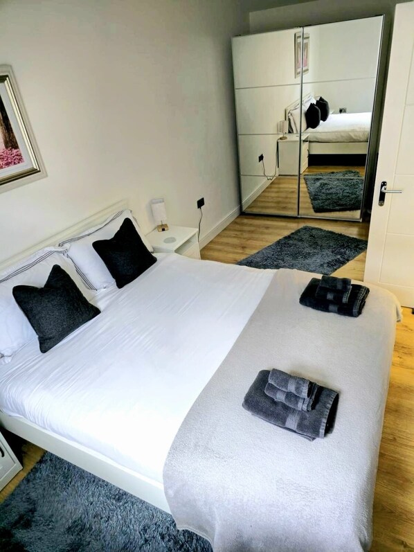 Una cama bien hecha con sábanas blancas y dos almohadas oscuras, una mesita de noche con lámpara, y un espejo grande que refleja la habitación.