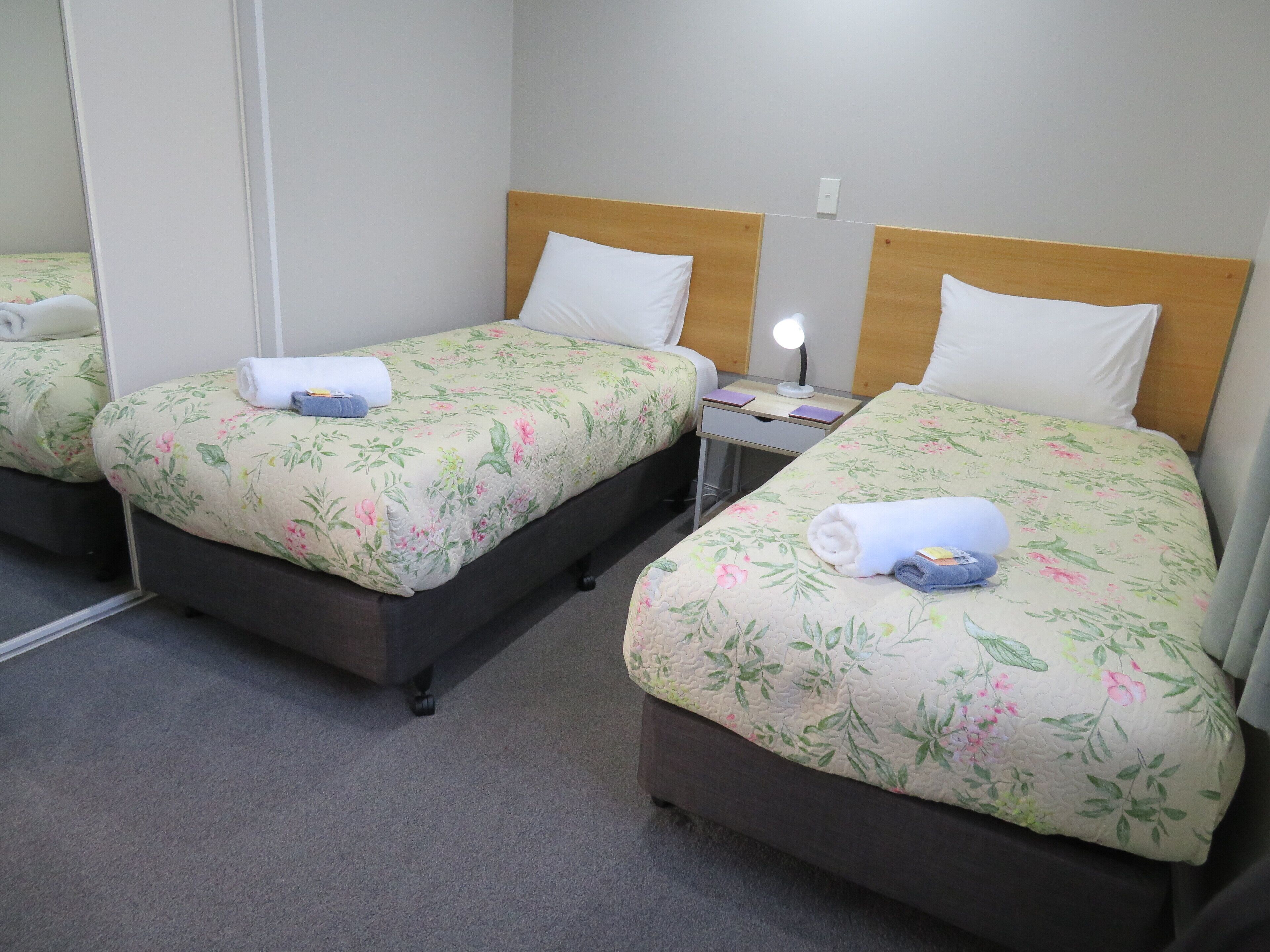 Una habitación de hotel con dos camas individuales, cada una con ropa de cama floral y un cabecero de madera. Hay una mesita de noche con una lámpara entre las camas.