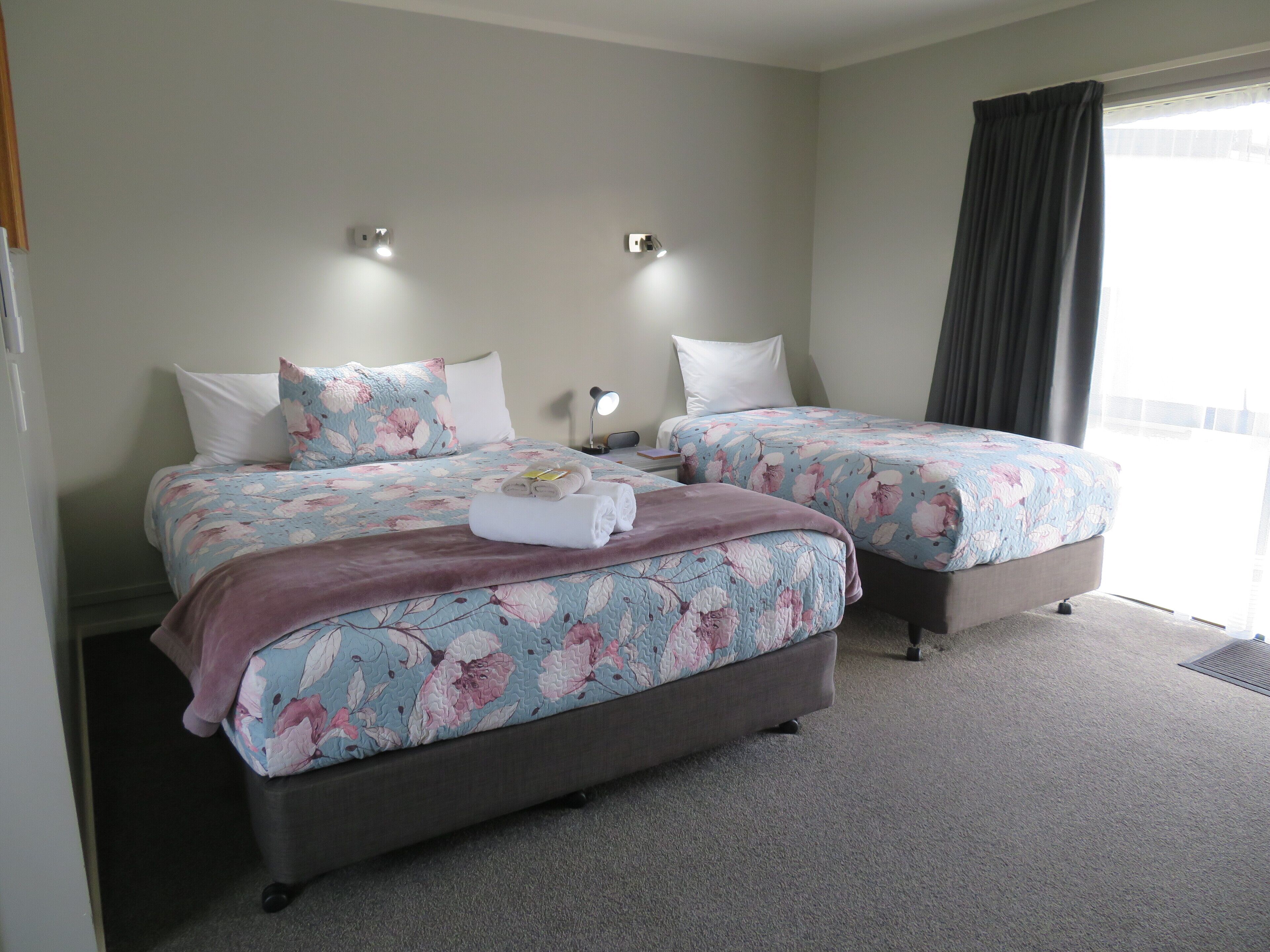A hotel room with two beds, floral bedding, a dark-colored sofa, a nightstand with a lamp, and a large window with curtains.