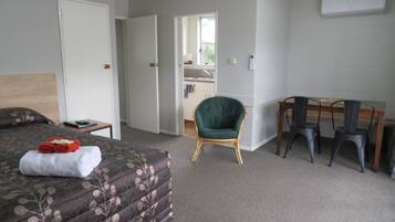 A hotel room with a bed, a desk, two chairs, a green armchair, and a kitchenette in the background.