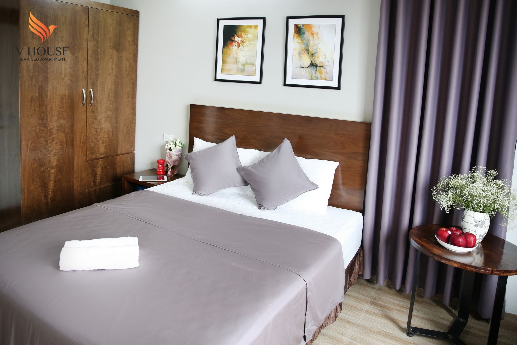 A neatly made bed with a grey bedspread and pillows, a wooden wardrobe, two framed artworks on the wall, and a small table with a vase and apples.