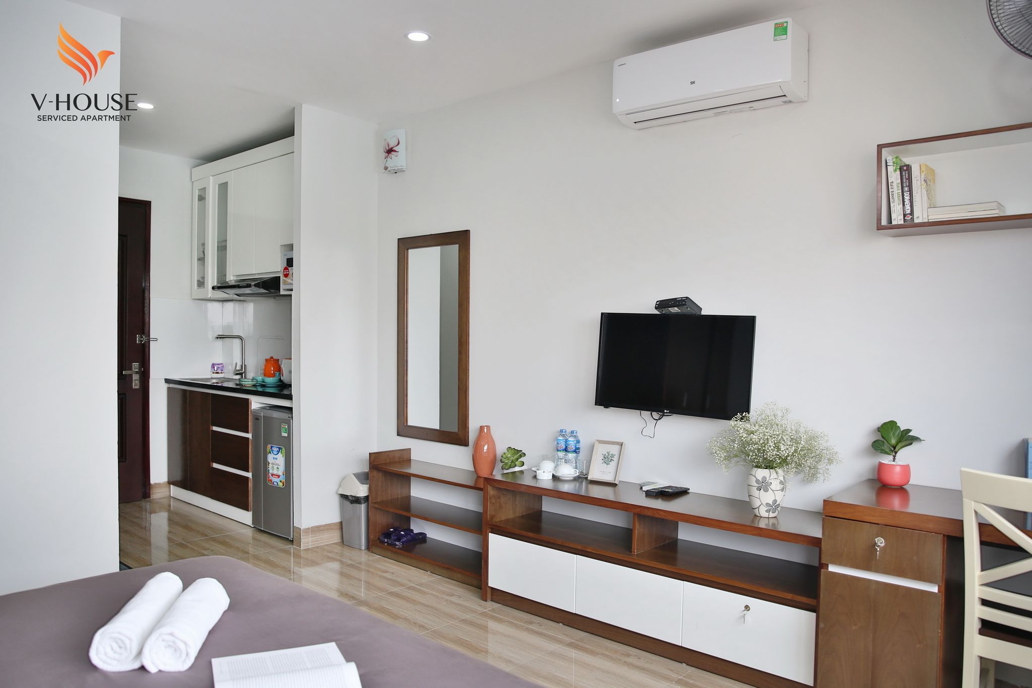 A serviced apartment with a kitchenette, a TV mounted on the wall, a wooden console table, and a bed with rolled towels.