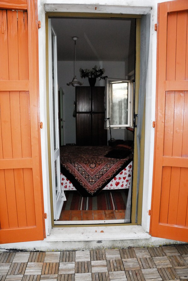 A room with an open door, a bed with a patterned bedspread, a hanging light fixture, and a window with a view of the outside.