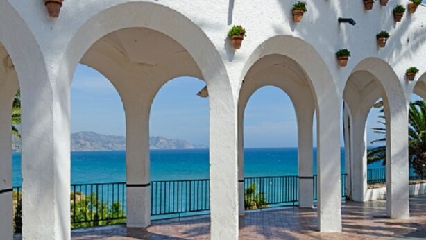 Une série d’arches blanches ornées de plantes en pot, qui mènent à un point de vue sur la mer.