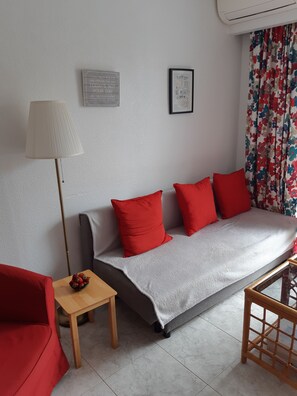 Un salón acogedor con un sofá, cojines rojos, una mesita con un cuenco de fresas, una lámpara de pie y una cita enmarcada en la pared.