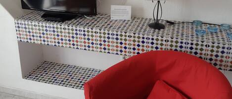 Une pièce dotée d’un téléviseur à écran plat, d’un fauteuil rouge, d’une table à motifs et d’une lampe fixée au mur.