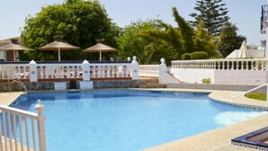 Une piscine entourée d’une clôture blanche, bordée d’arbres, avec un bâtiment blanc en arrière-plan.
