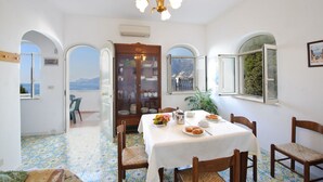 Un'area pranzo con tavolo apparecchiato per la colazione, un mobile contenitore in legno con vetrate e vista sul mare attraverso una finestra ad arco.