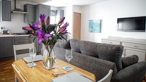 A modern living room with a grey sofa, a wooden dining table set with cutlery and glasses, and a vase with purple flowers. - Cottage sleeps 4 with private hot tub. Edinburgh festival less than 1hr away.  (Kinross)