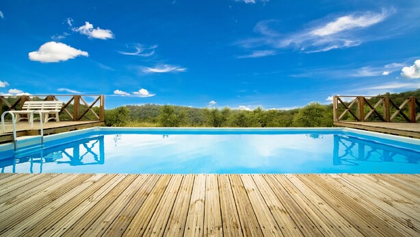 Une terrasse en bois avec une piscine à l’eau cristalline, des bancs en bois et une vue sur une zone boisée sous un ciel bleu.