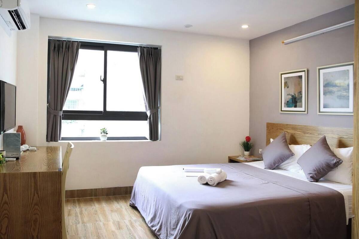 A hotel room with a bed, a desk, a window with curtains, a nightstand, and two framed pictures on the wall.