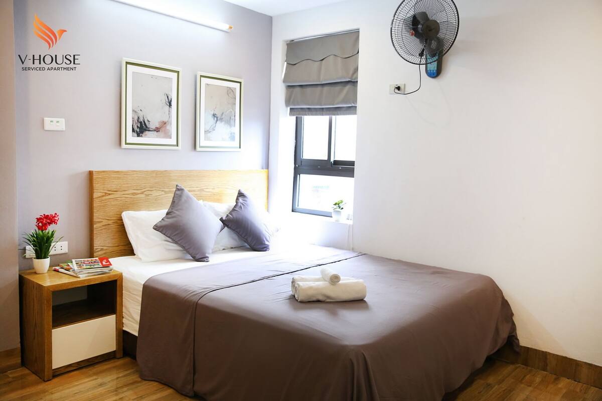 A bedroom with a wooden headboard, a brown bedspread, a white pillow, a grey pillow, a small bedside table with a plant and a magazine, and a wall-mounted fan.