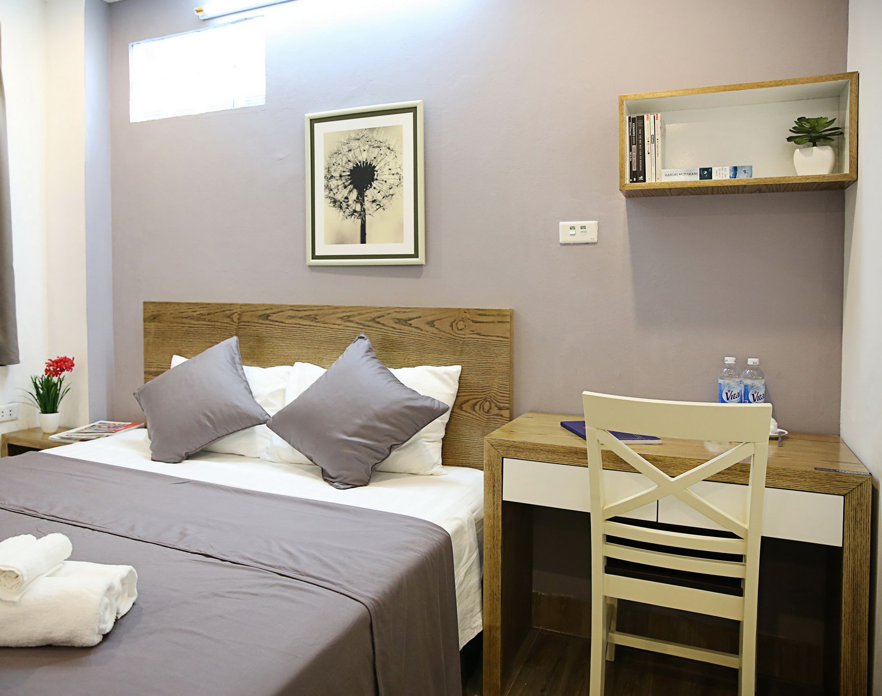 Una camera d'hotel con un letto, una scrivania, una sedia, un piccolo ripiano con libri e una pianta, e un quadro appeso al muro.