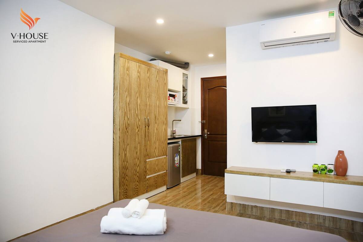 A serviced apartment with a wooden wardrobe, a kitchenette, a TV mounted on the wall, and a small table with a towel.