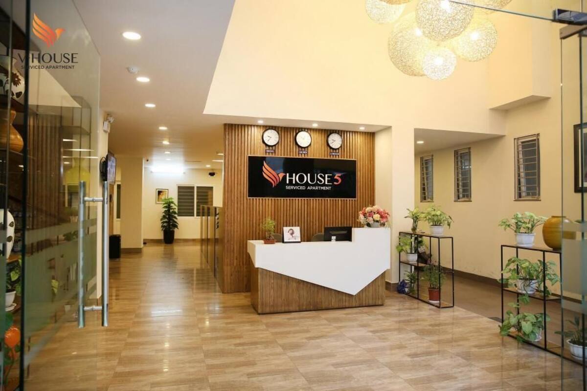 A reception desk with a sign for 'HOUSE 5 Serviced Apartment,' a wooden reception desk, a wall-mounted clock, and potted plants.