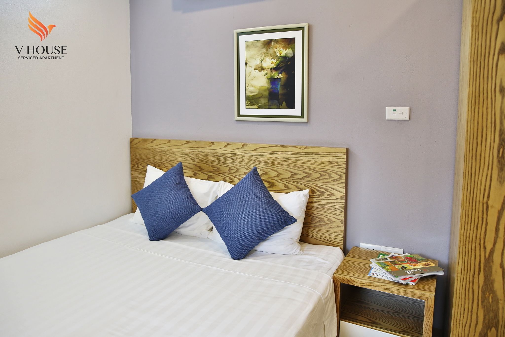 Una cama bien hecha con almohadas azules y blancas, una obra de arte enmarcada en la pared y una mesita de noche de madera con revistas.