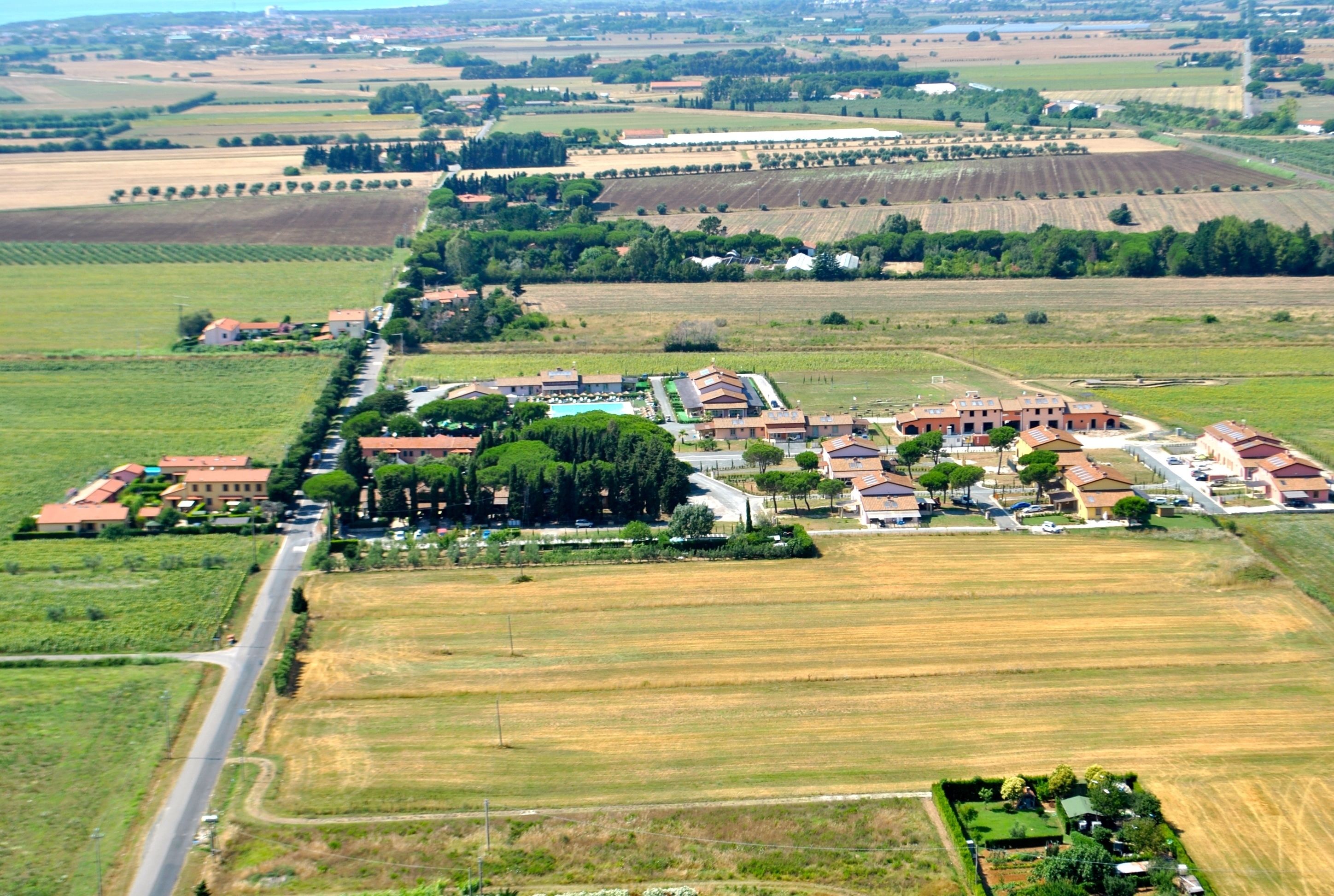 An aerial view of a rural area with a cluster of buildings, including a large house with a swimming pool, surrounded by fields and a road.