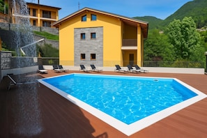 A swimming pool with lounge chairs, a stone wall, and a yellow building with a balcony.